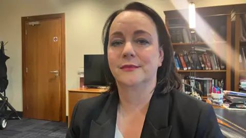 Catherine Kierans sits in an office, books are behind her in a bookcase and a brown wooden door is to her right. She has shoulder-length brown hair and wears red lipstick. She has a black jacket on.