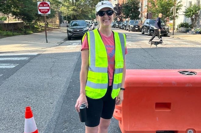 A white woman with a beige hat, dark sunglasses, a red t-shirt, a neon vest with reflective strips and black shorts stands in the street, holding a drink, next to an orange barricade.