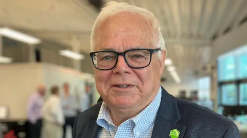 BBC Steve Berry, an older man with short, white hair, smiles at the camera. He is wearing black-framed square glasses, a grey suit jacket and a blue and white checkered shirt. He is also wearing a green pin. He is standing in a large room with people in the background, but they are blurred.