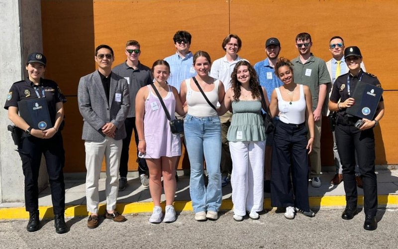 Group photo of a diverse team of young adults and two police officers, all smiling and standing together outdoors.