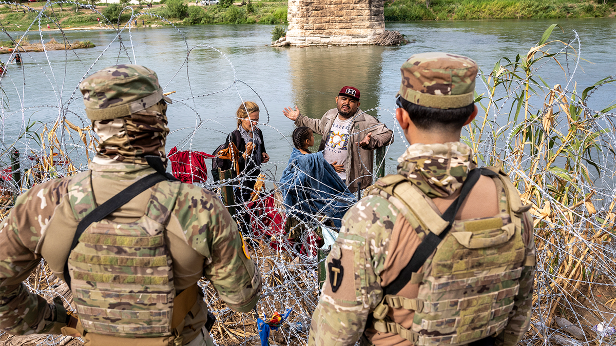 migrants wade through river too razor wire while men in camouflage fatigues look on
