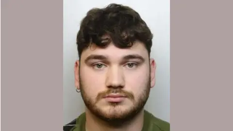 Avon and Somerset Police George Rogers looking into the camera for a police custody image with his left ear pierced, short dark brown curly hair and a beard