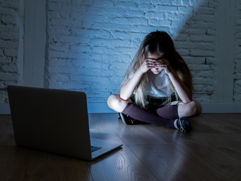 A sad girl holds her head in dismay while sitting opposite her laptop in a dark room