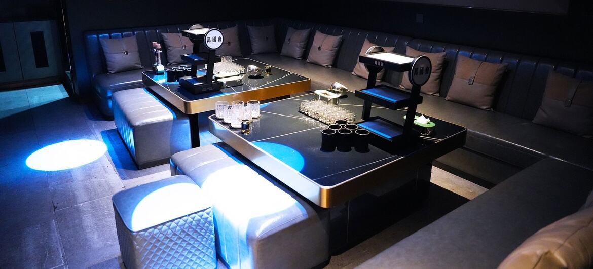 A modern, dimly lit private karaoke room with plush grey sofas arranged around glossy black tables, illuminated by blue and white LED lights.