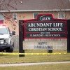 This photo shows a large wooden sign that says "Abundant Life Christian School" on it. An emergency vehicle is parked on the grass next to the sign. Yellow caution tape extends across the photo.