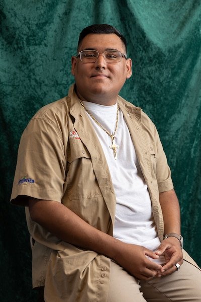 A young brown man woman with short dark hair and wearing a tan button up shirt with a white undershirt and gold chains with a cross poses for a portrait in front of a velvet green backdrop.