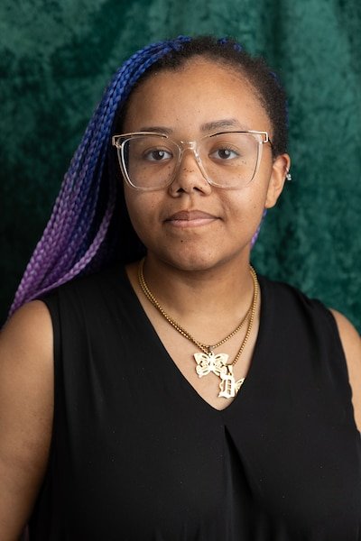 A young Black woman with long colorful braids and wearing a black blouse poses for a portrait in front of a velvet green backdrop.