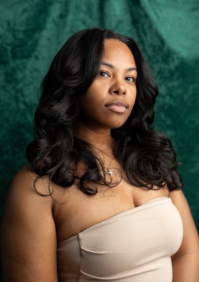 A young Black woman with long dark hair and wearing a tan tube top poses for a portrait in front of a velvet green backdrop.