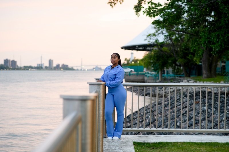 A young Black woman wearing a baby blue  jumpsuit poses for a portrait in front of the Detroit skyline and the Detroit River.
