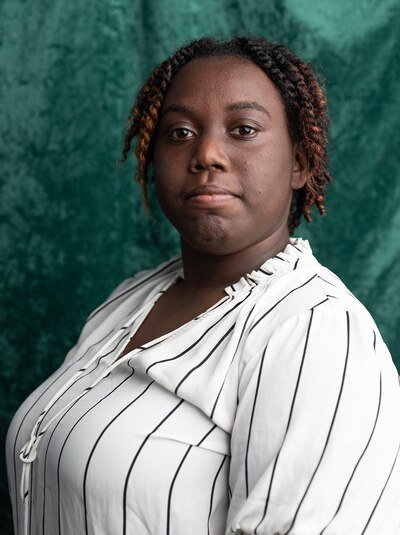 A young Black woman with short dark hair and wearing a white and black blouse poses for a portrait in front of a velvet green backdrop.
