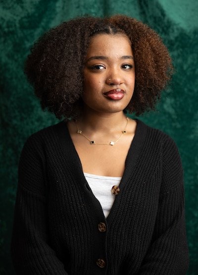 A young Black woman with dark short hair and wearing a black sweater with a white under shirt poses for a portrait in front of a velvet green backdrop.